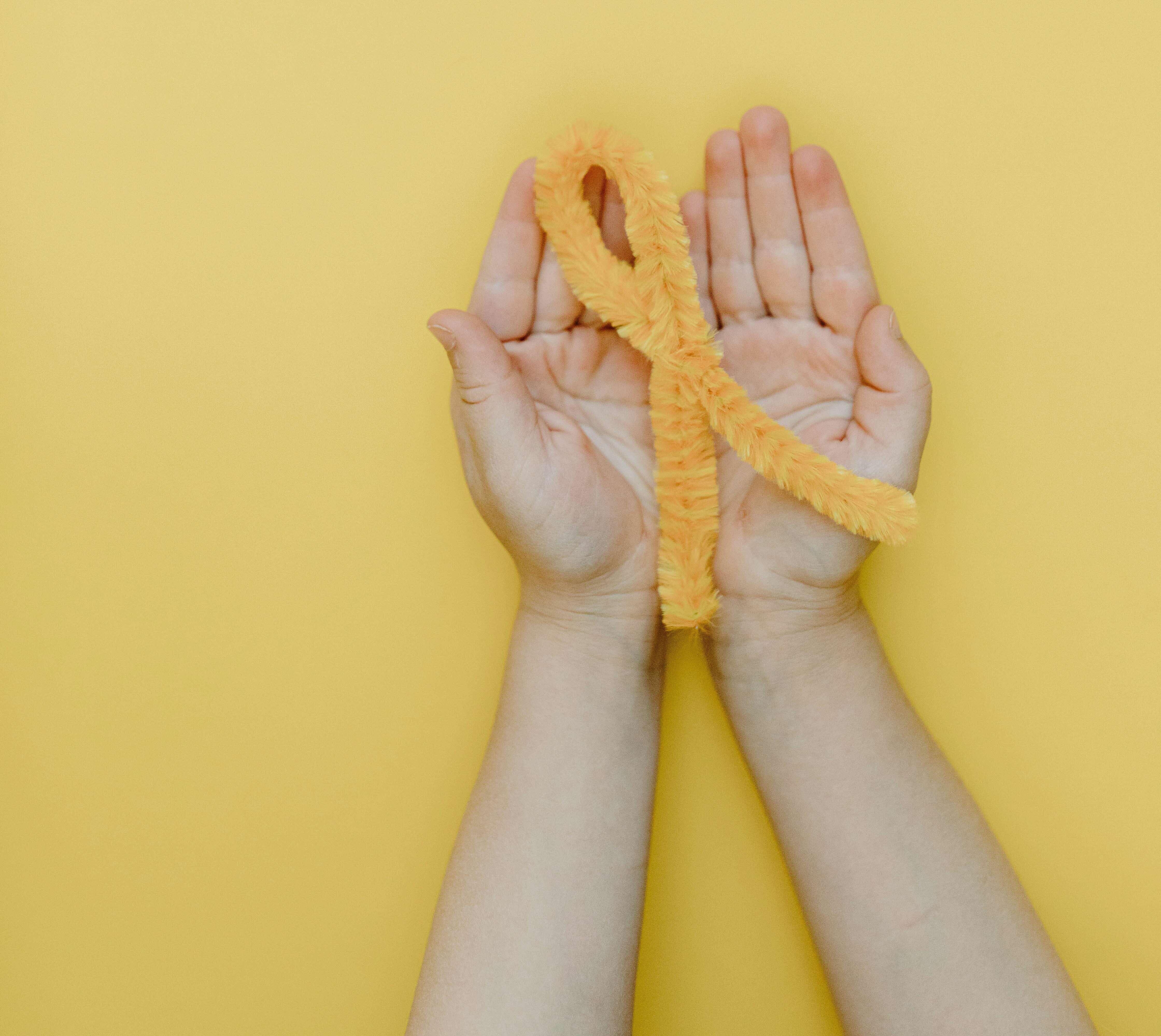 Hands holding a fuzzy yellow awareness ribbon, centered on a yellow background.